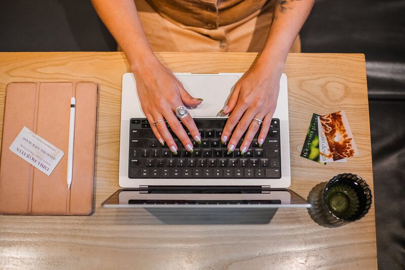 A computer sits on a bed next to a notebook, phone, and pillows