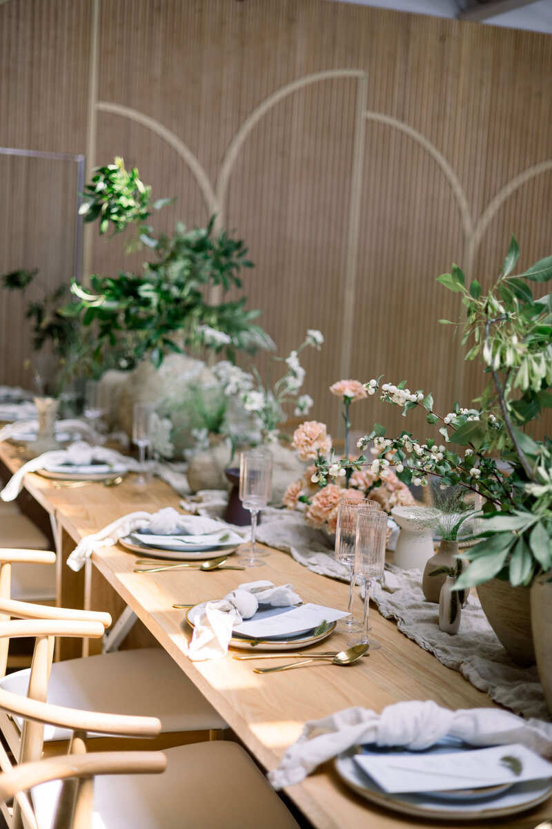 Wood banquet table styled with potted greenery, neutral ceramics, gauze runner, and natural wishbone chairs in a modern space.