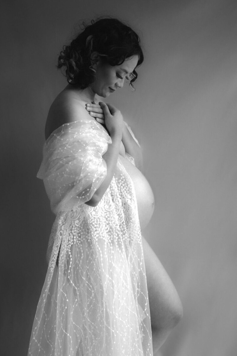 Expectant mother draped in delicate lace standing in profile with hands crossed over her chest and gently smiling at her baby bump.