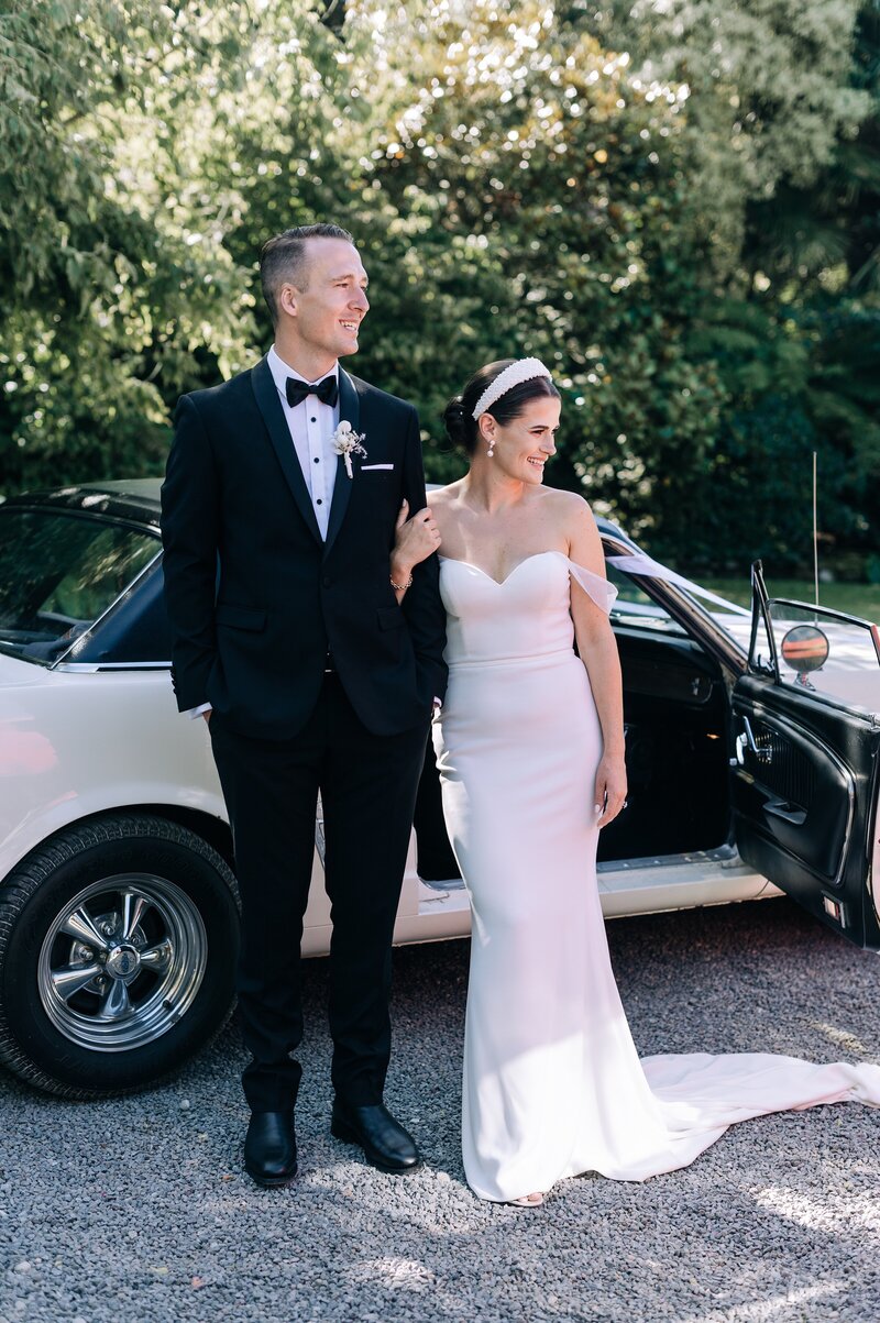 bride in trish peng wedding dress and groom in black tuxedo in front of ivory mustang at their christchurch backyard wedding