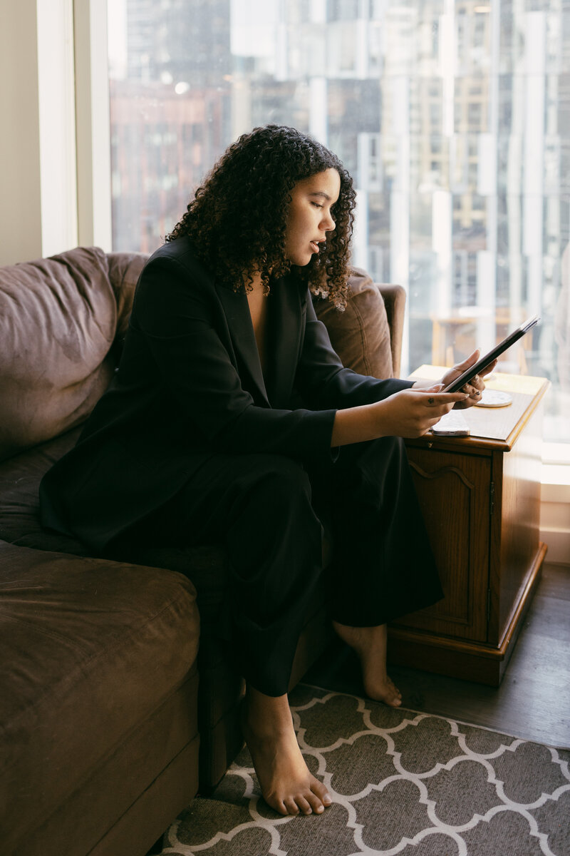 Catryce scrolling on a tablet while sitting on a couch in a pantsuit