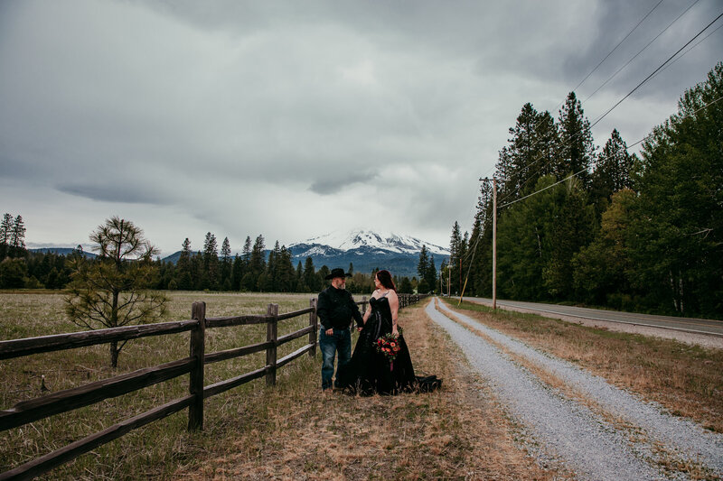 Bride and groom standing near a rustic fence during their Mount Shasta elopement, with dramatic clouds, open fields, and the snow-covered mountain in the distance.