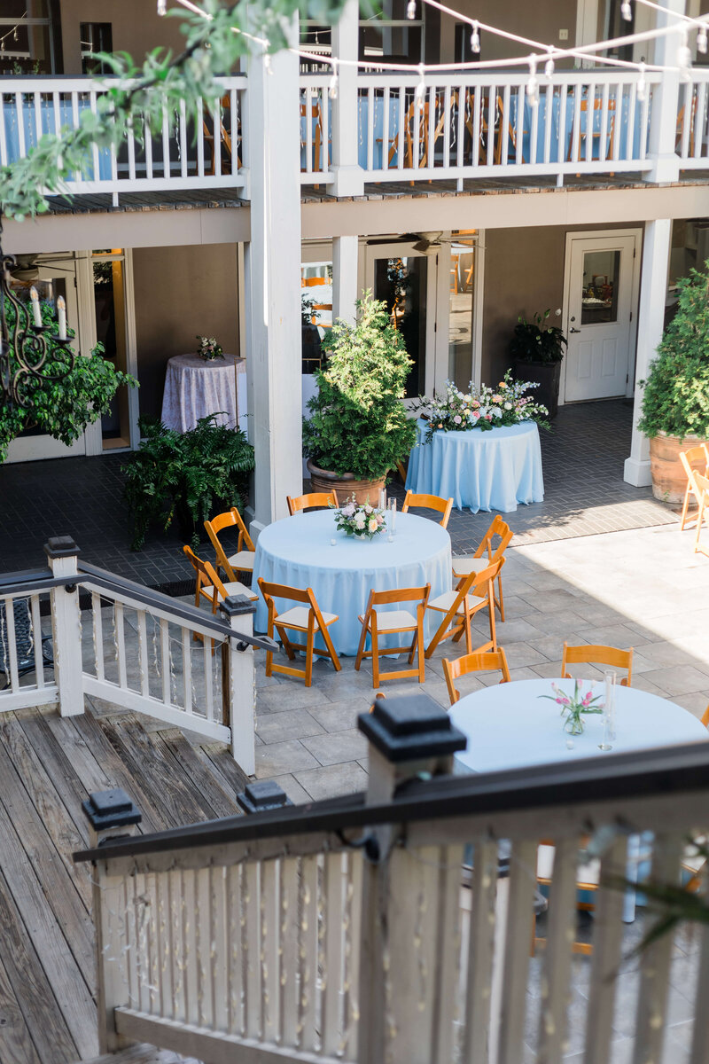 An outdoor courtyard set for an event, with round tables covered in white cloths, wooden chairs, floral centerpieces, potted plants, and string lights overhead.