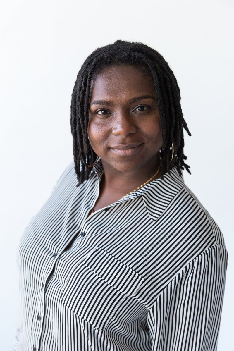 A woman with shoulder-length dreadlocks wears a black-and-white striped blouse against a clean, white background. A striking and professional headshot.
