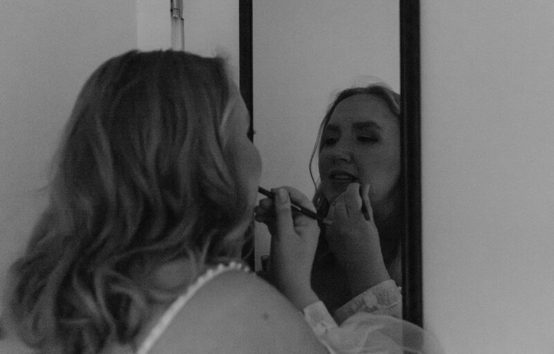 black and white photo of a bride applying lipstick in front of a mirror, captured during a quiet and personal getting-ready moment before the ceremony. Her reflection shows a calm, focused expression as she carefully enhances her makeup, wearing a gown with delicate lace straps.