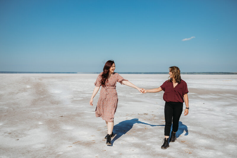 Two women walking hand in hand across a vast, salt flat under a clear blue sky