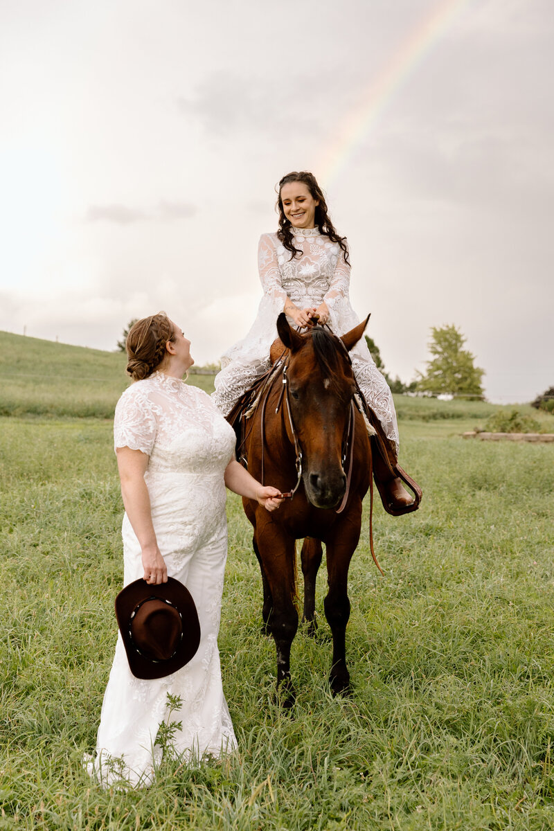 LGBTQ+ couple eloping at a horse farm, one bride is riding the horse while the other bride is holding the reins. There is a rainbow behind them.