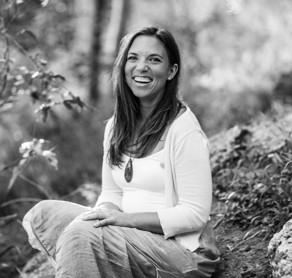 Women in golden light, wearing dark rimmed glasses and striped shirt, smiling down at her hands as she rolls out clay