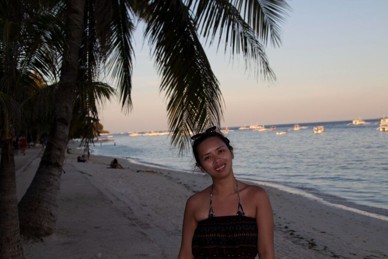 Young woman smiling on a beach walkway at sunset, standing under palm leaves with the ocean and boats in the background