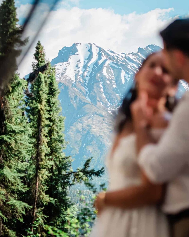 A bride and groom lean into each other, about to kiss during their luxury Alaska elopement on the Matanuska glacier.  Behind them, the tall, snow covered mountain fills the frame with peeks of bright blue sky.