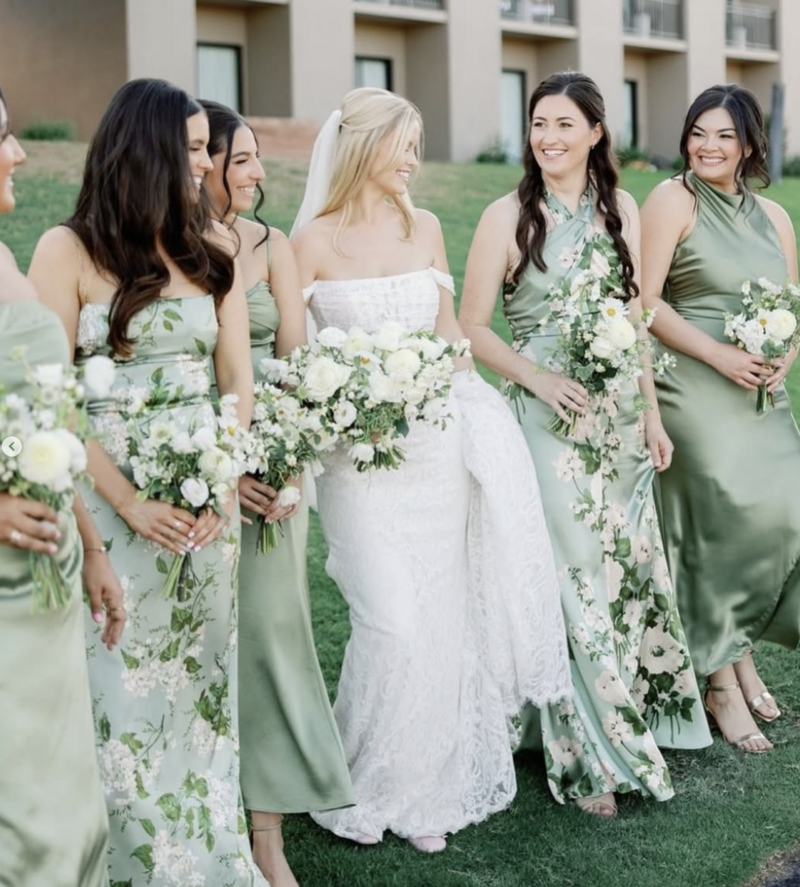 Bride and her bridesmaids laughing and smiling, all walking and looking at one another. Bride is wearing a lace fitted Justin Alexander wedding dress and bridesmaids are all in floral green dresses with flower bouquets.