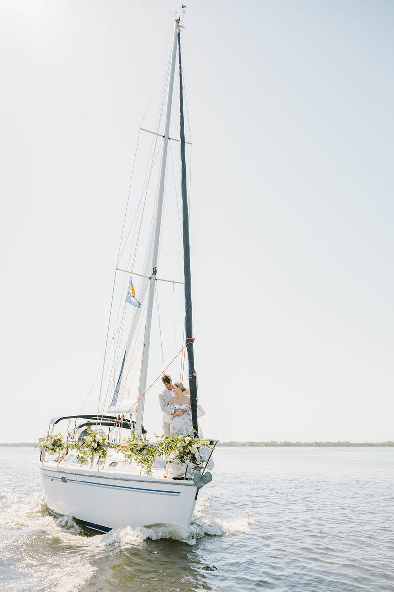 Newport RI Wedding Photographer | A sailboat glides on calm waters under a clear sky, decorated with green foliage. Two people stand at the front, creating a serene, romantic scene.