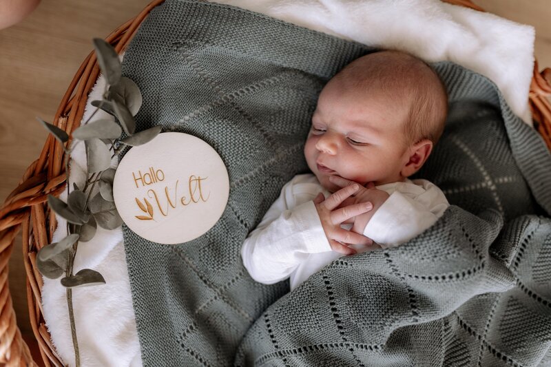 Baby liegt auf einer gruenen Decke in einem Korb und hat Hallo Welt neben sich stehen