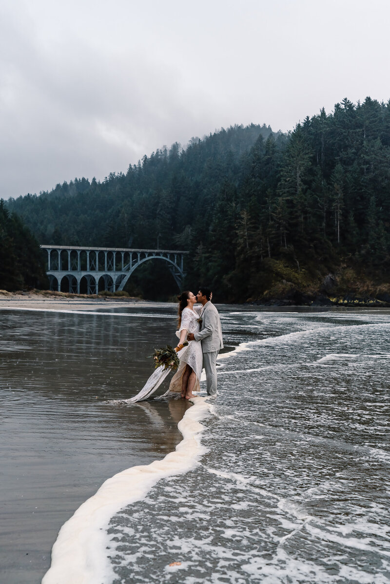 During their moody oregon coast elopement, a bride and groom kiss on the beach in their wedding attire. The waves roll in between them.