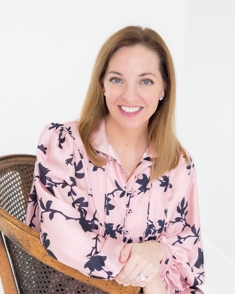 family travel expert for international travel headshot with woman in a pink and dark blue dress posing on a rattan chair on a white backdrop