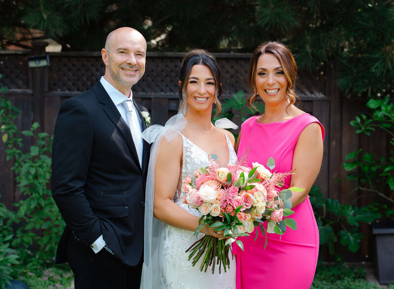 a bride smiling and standing between her mother and father on wedding day.  Captured outdoors by Ottawa wedding photographer JEMMAN Photography