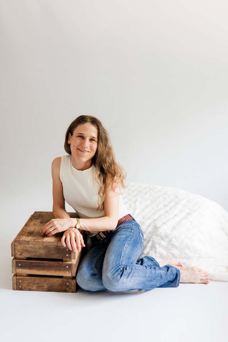 Woman sitting down and leaning against a brown wooden crate, wearing faded blue jeans and cream top while smiling and looking happy.