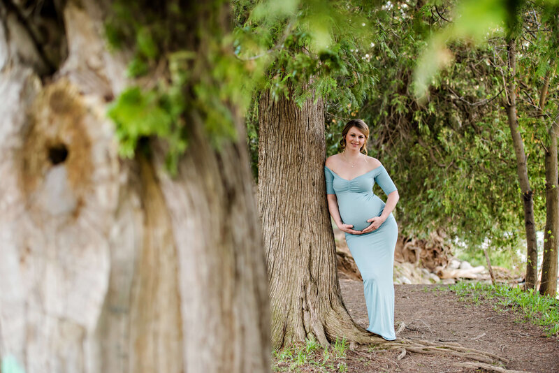 Pregnant woman wearing a blue maternity dress leaning against a tree holding her belly for her outdoor Burlington, ON maternity photography session.
