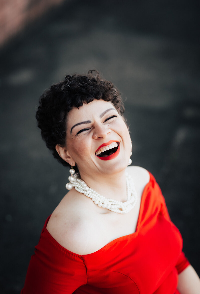 The camera frames Laura's head and shoulders from above, as she laughs with an open smile and closed eyes toward the camera. She is wearing a red, sweatheart neckline dress, with multiple strings of white pearls around her neck.