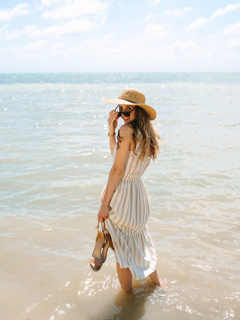 Kilee in an orange dress and straw hat sitting on a palm tree with a book in front of the ocean