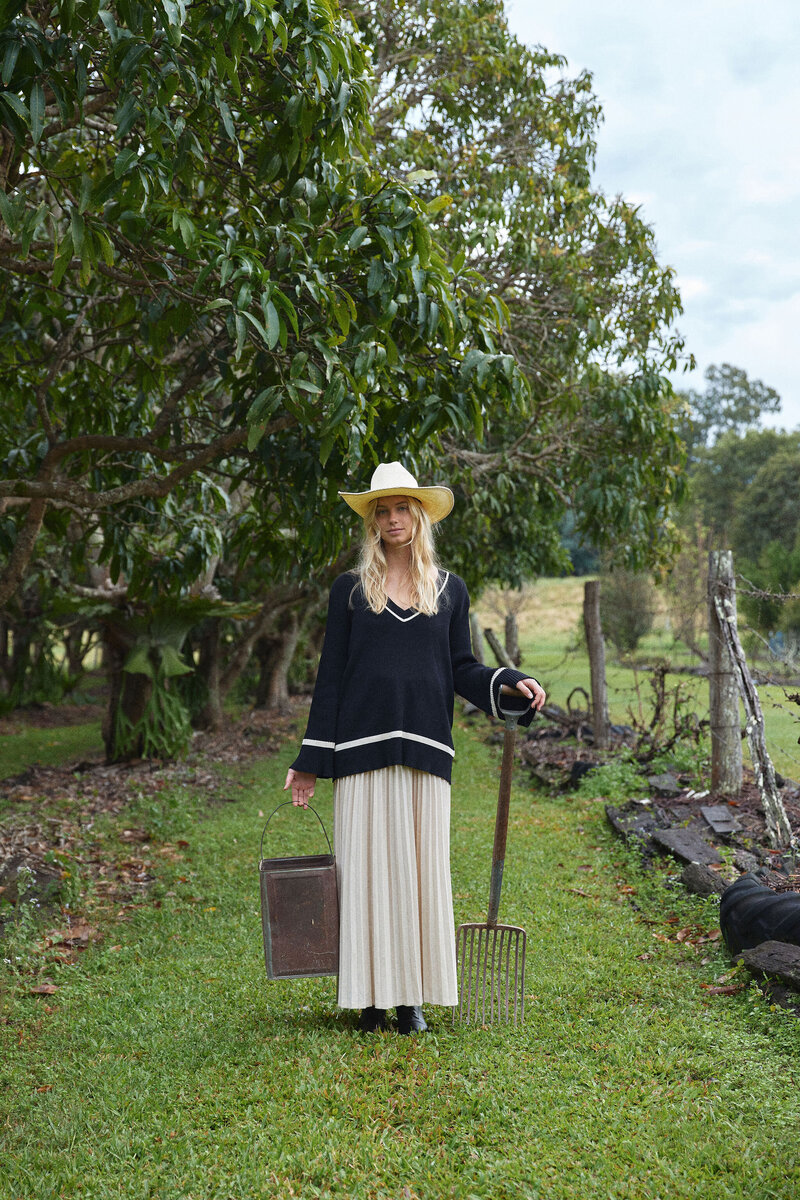 Model standing with rustic rake and water can wearing long white dress and oversized knit