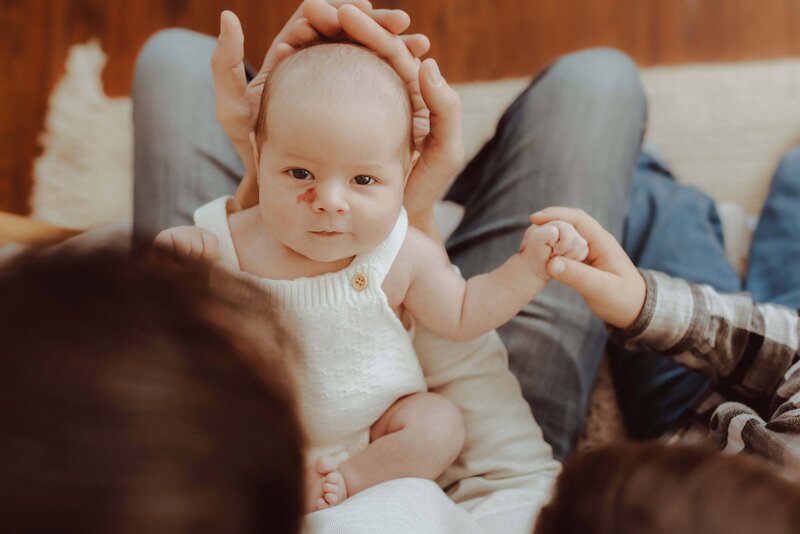 Close-up of a baby in a white outfit, held by an adult, reaching out towards a child's hand. The baby is sitting on a person's lap.