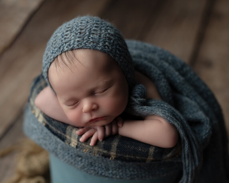 Newborn boy posed in a blue knit bonnet and wrap in a rustic bucket on wood floors—styled with warmth and intention