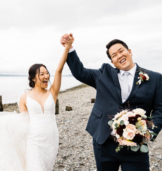 A bride and groom raise their hands in the air in celebration after their Pacific Northwest elopement on the beach in north Seattle