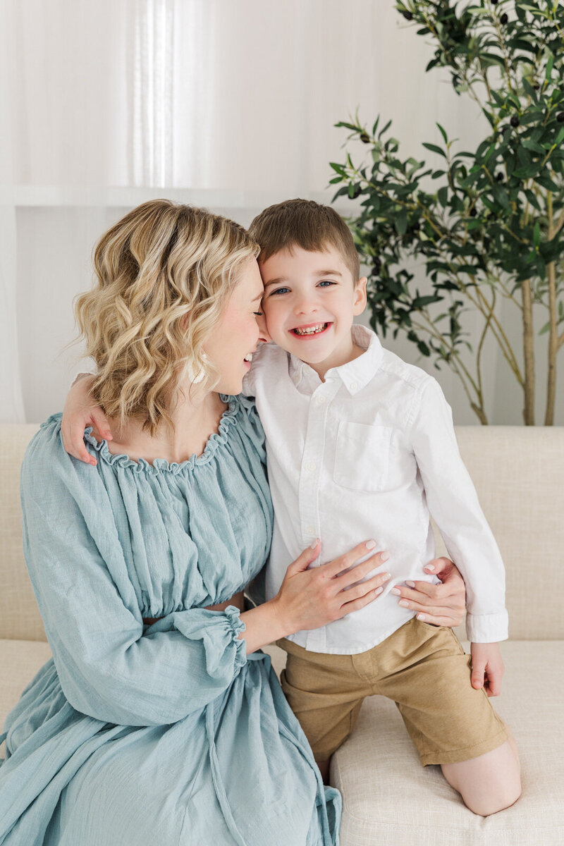 Young boy smiles as mom snuggles him during family session