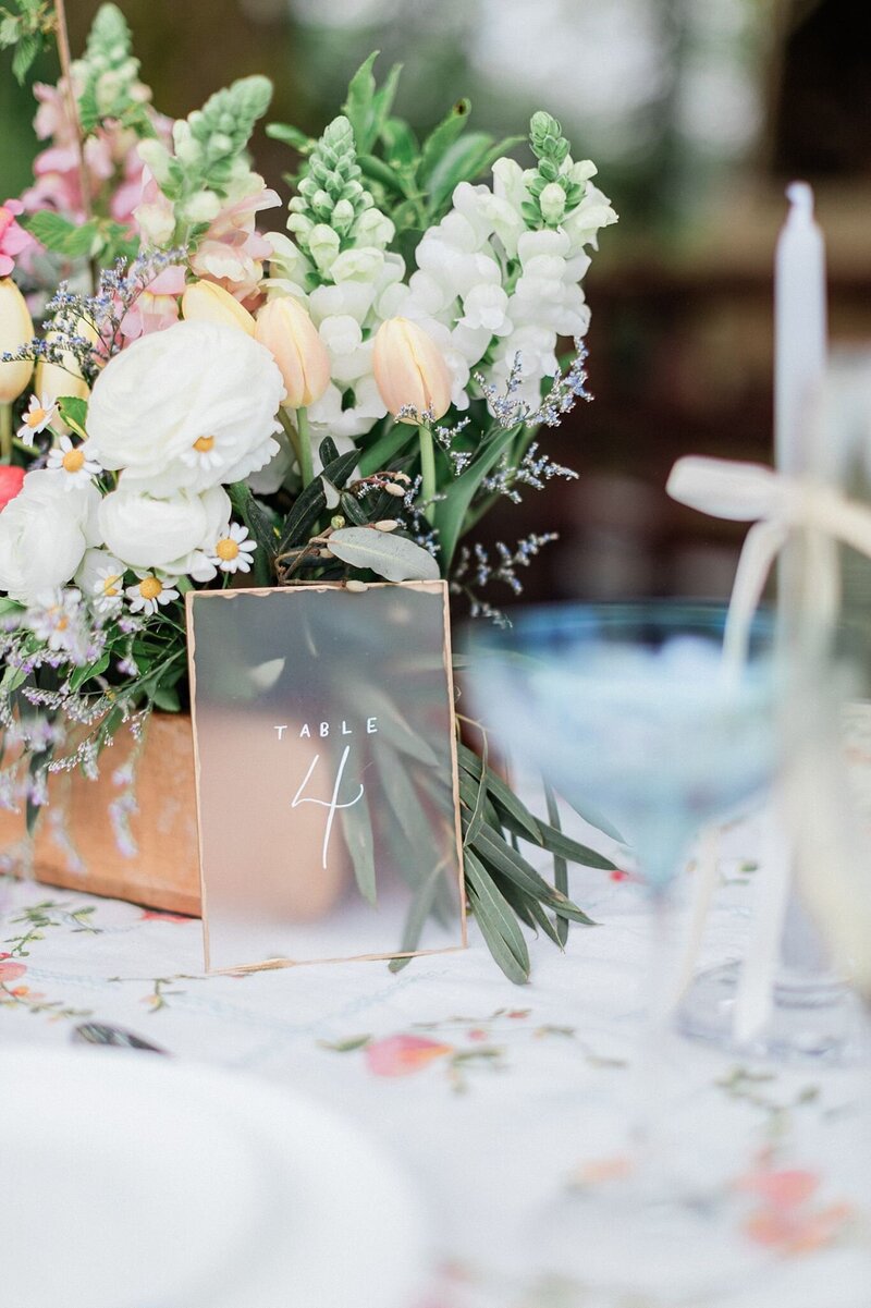 Wedding table number sign in acrylic with handwritten script, styled next to a pastel floral arrangement featuring tulips, ranunculus, and olive branches