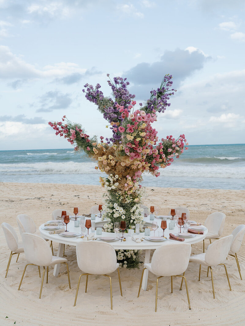 White and gold chairs surround a round white wedding reception table with a towering floral centerpiece on a Mexico beach