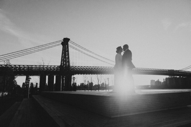 Black and white of bride and groom standing in front of bridge at sunset