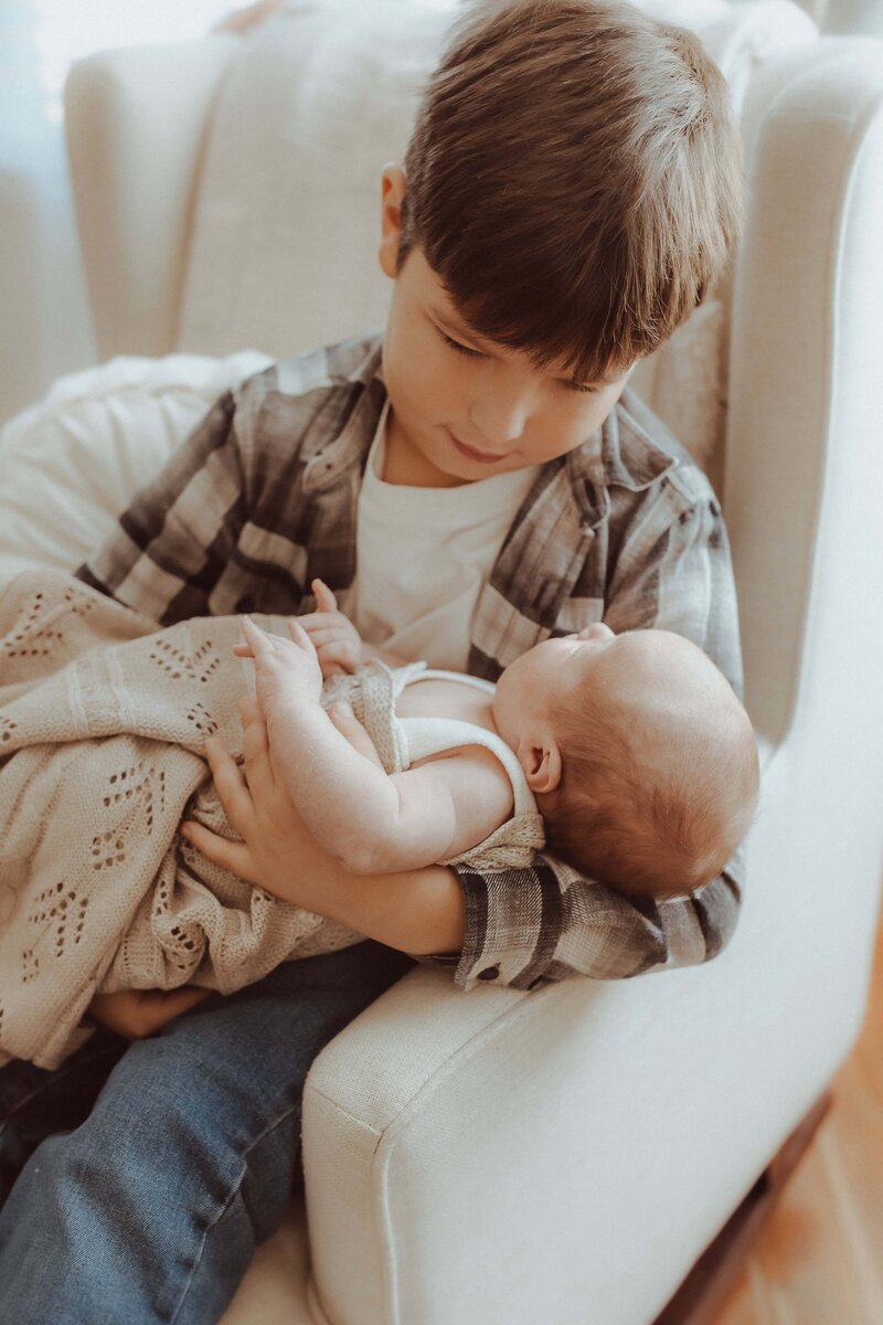 A young boy in a plaid shirt sits on a chair, gently holding a baby wrapped in a knitted blanket.