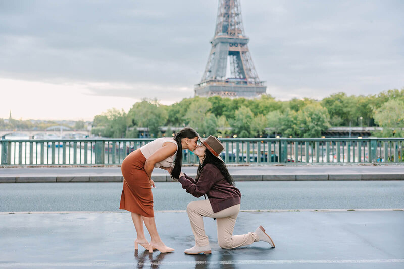 Lesbian couple kissing in front of the Eiffel Tower at Bir-Hakeim Bridge, with one woman kneeling after a surprise proposal during a romantic Paris photoshoot.