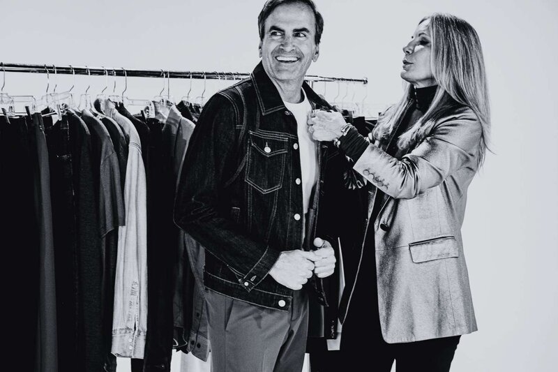 black and white image of a man smiling while a woman adjusts his denim jacket during a styling session with clothing racks in the background
