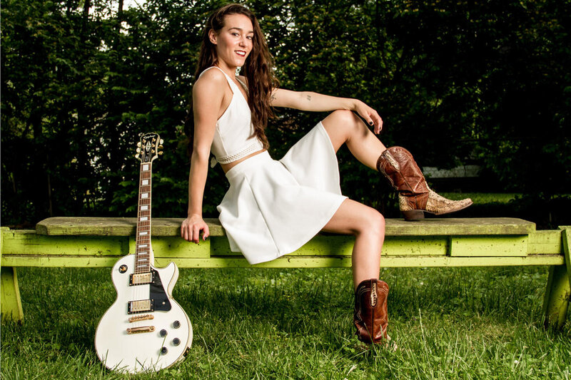Country Music portrait Nashville Leanne Pearson sitting on bench in white dress with cowboy boots white electric guitar leaning beside her  trees behind her grass below