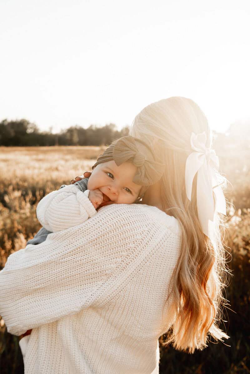 Baby in a bow headwrap snuggled on her mother’s shoulder during a golden hour motherhood session in a sunlit Florida field.