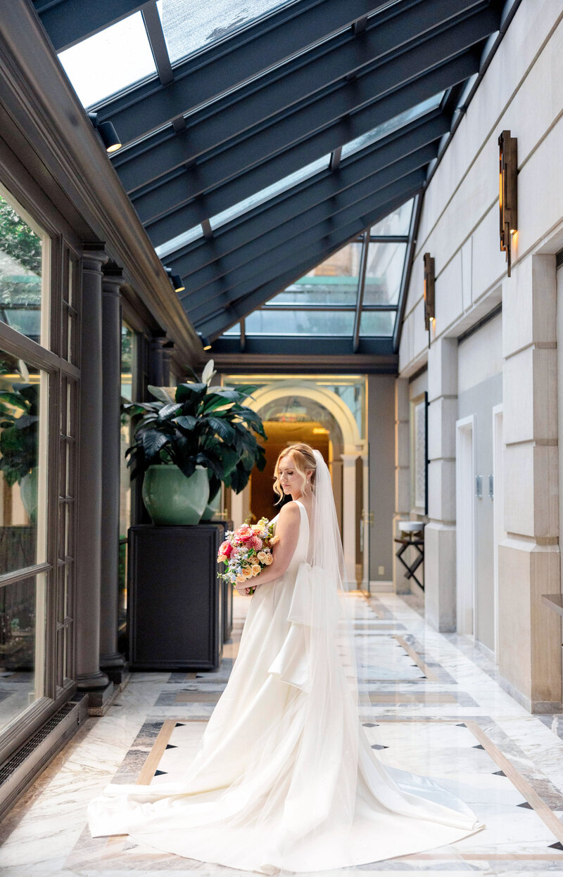 A bride in a long white dress and veil stands in a bright glass-ceiling hallway, holding a colorful bouquet. Elegant marble floors and large potted plants surround her, with natural light illuminating the scene.