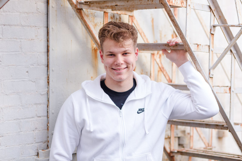 waynedale high school senior guy standing under rusted metal steps wearing a white nike long sleeve shirt, one arm holding onto a step, photographed Downtown Wooster Ohio, photographed by Jamie Lynette Photography canton ohio senior photographer