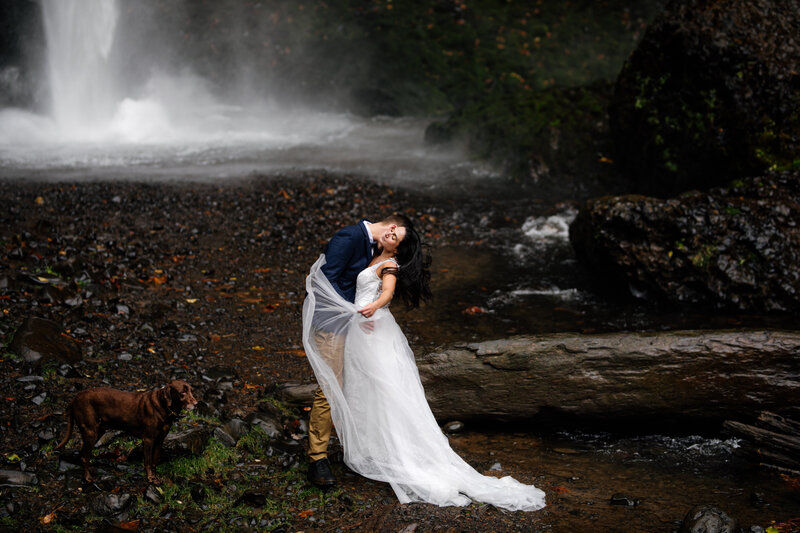couple kissing in front of foggy falls A bride and groom embrace on a cliff with the Oregon coast in the background, under a dramatic sunset, photographed by an Oregon elopement photographer.