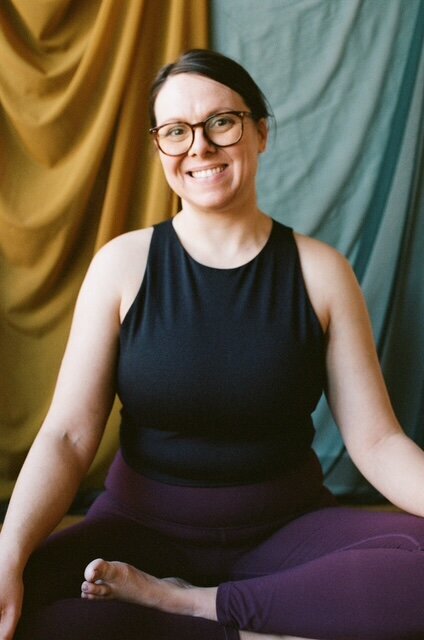 A woman wearing glasses and a black top is seated in a yoga pose, demonstrating focus and tranquility.