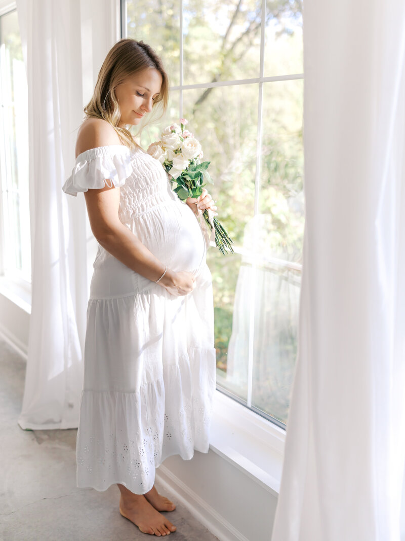 Woman in white dress holding pregnant belly in a minneapolis studio for her minneapolis maternity photography session with Angela Watts Photography.