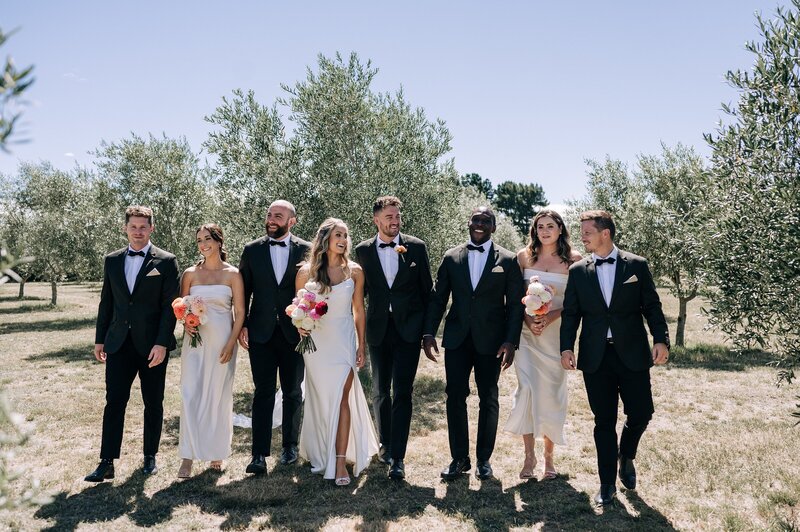 a bridal party in black and white on a sunny day in a waipara olive grove with bright coloured dahlia bouquets