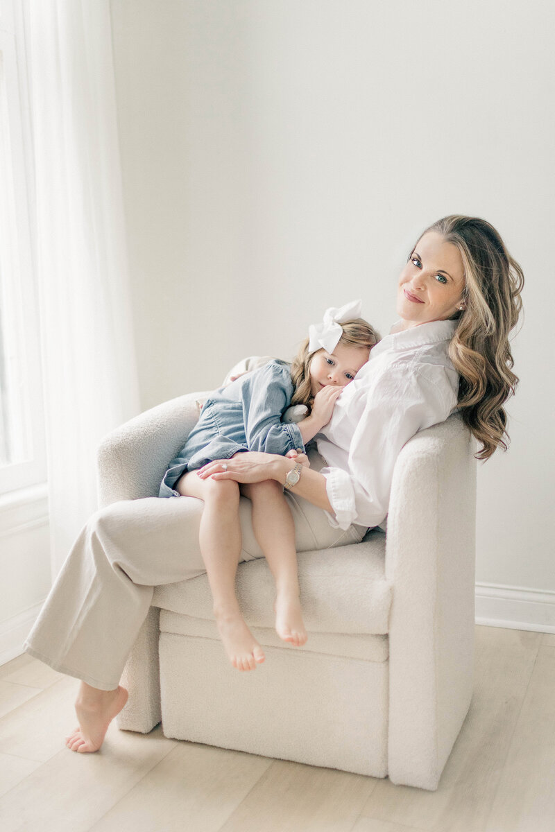 Mother and daughter cuddled together on a soft cream chair inside a light-filled studio during a Kansas City motherhood photography session.