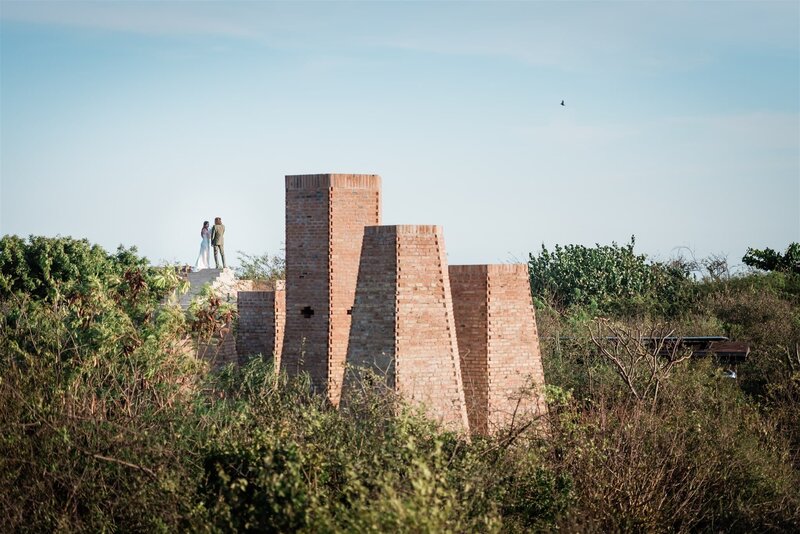 A couple in wedding finest stands atop a beauitiful brick viewing platform and neighboring spa building also of light red brick that rises out of the green plants during this adventure luxury elopement in Mexico.  They are far away from the photographer  so tiny in the beautiful frame. 