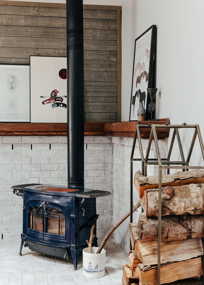 A blue wood burning stove with a stack of wood next to it and artwork on the mantle above it, representing a nordic interior design.