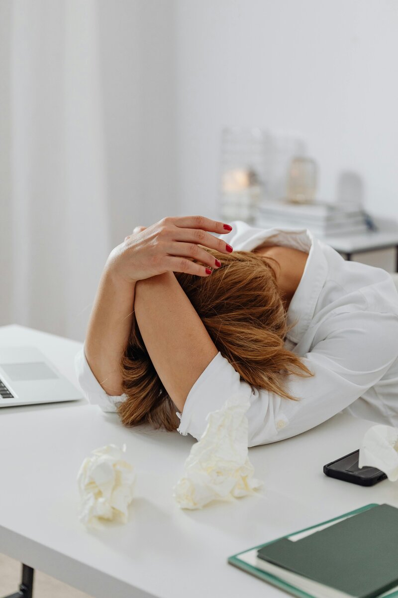 A woman struggling with intrusive thoughts, resting her head on a table with hands wrapped around her head in distress.