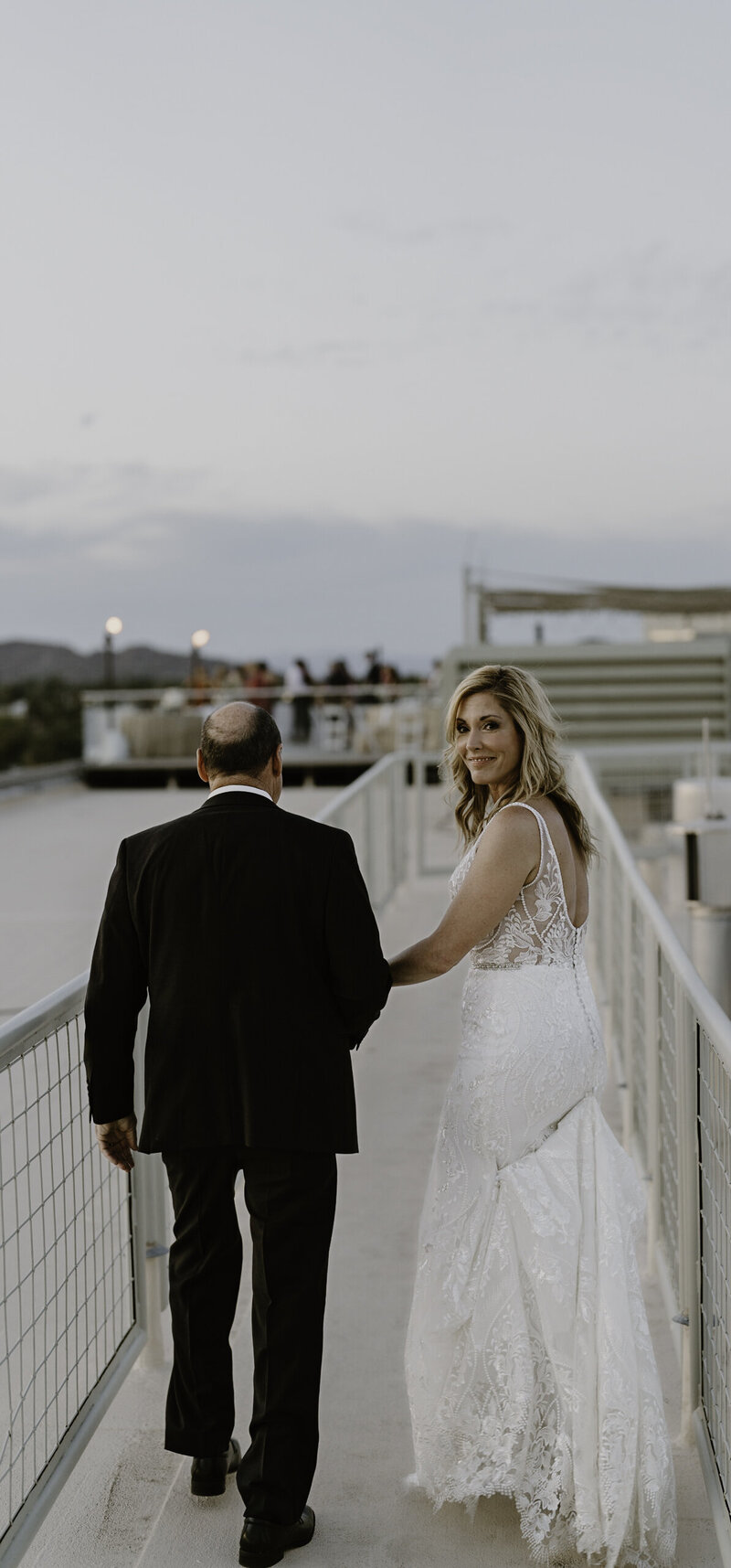 A bride in a detailed lace wedding gown walks along a bridge holding hands with a man in a black suit, looking back over her shoulder with a radiant smile. The bridge's railing and lighting create a modern and romantic evening atmosphere, with guests visible in the background enjoying the event.