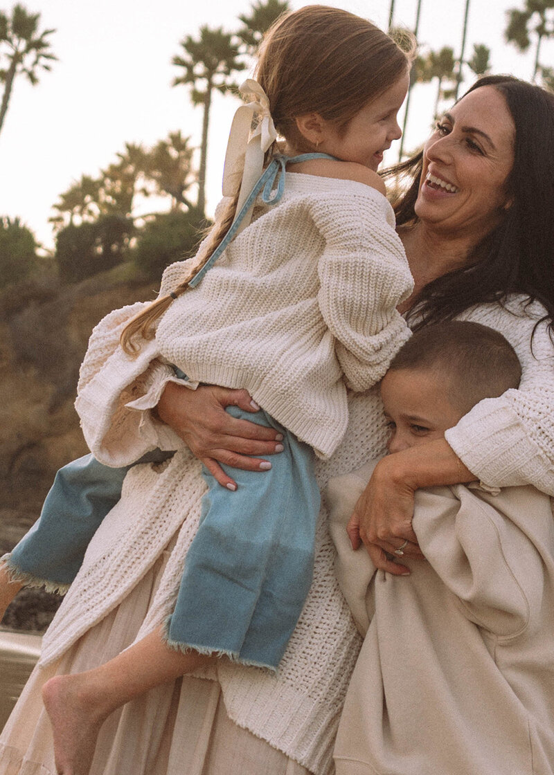 A candid close-up of a mother holding her daughter and laughing as her young son hugs her side, with soft sunset light and palm trees in the background at Laguna Beach.