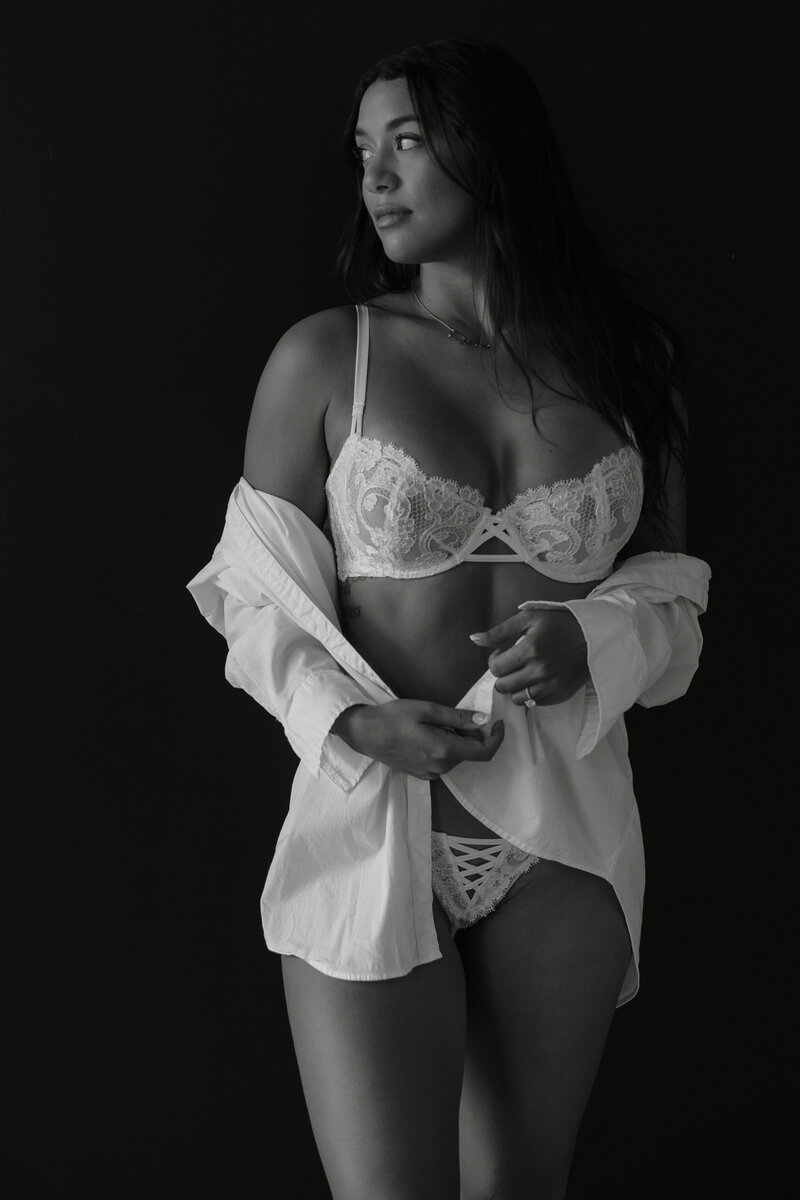 Black and white photo of woman in white lingerie with a white shirt around her arms standing in front of a black wall